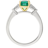 Oval emerald engagement ring showing yellow gold claw setting, diamond side stones and white metal band. Made in Hatton Garden